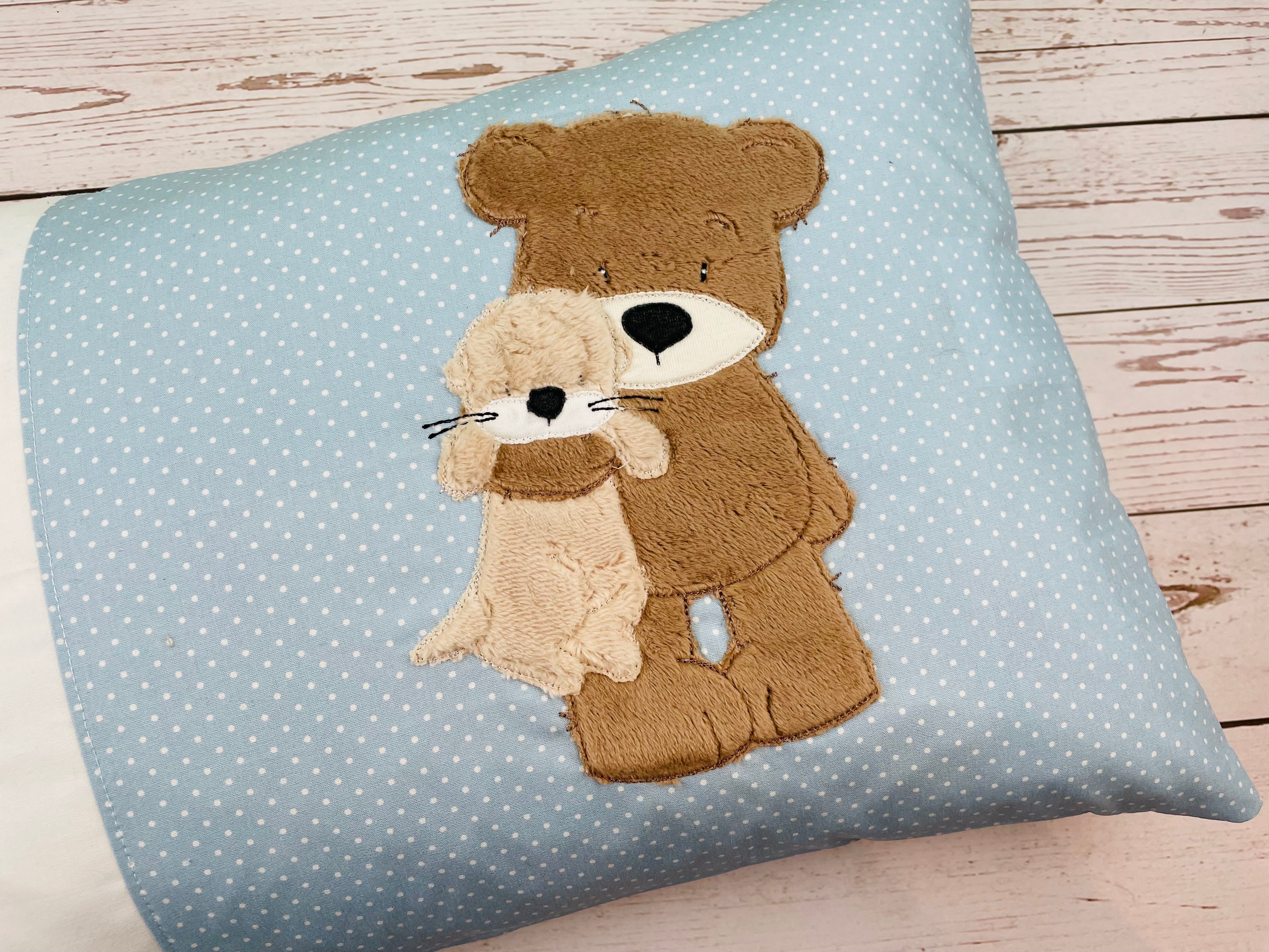 Kuschelkissen bestickt mit Teddybär der einen kleinen Otter im Arm hält. Personalisierbar mit Name und Geburtsdatum.