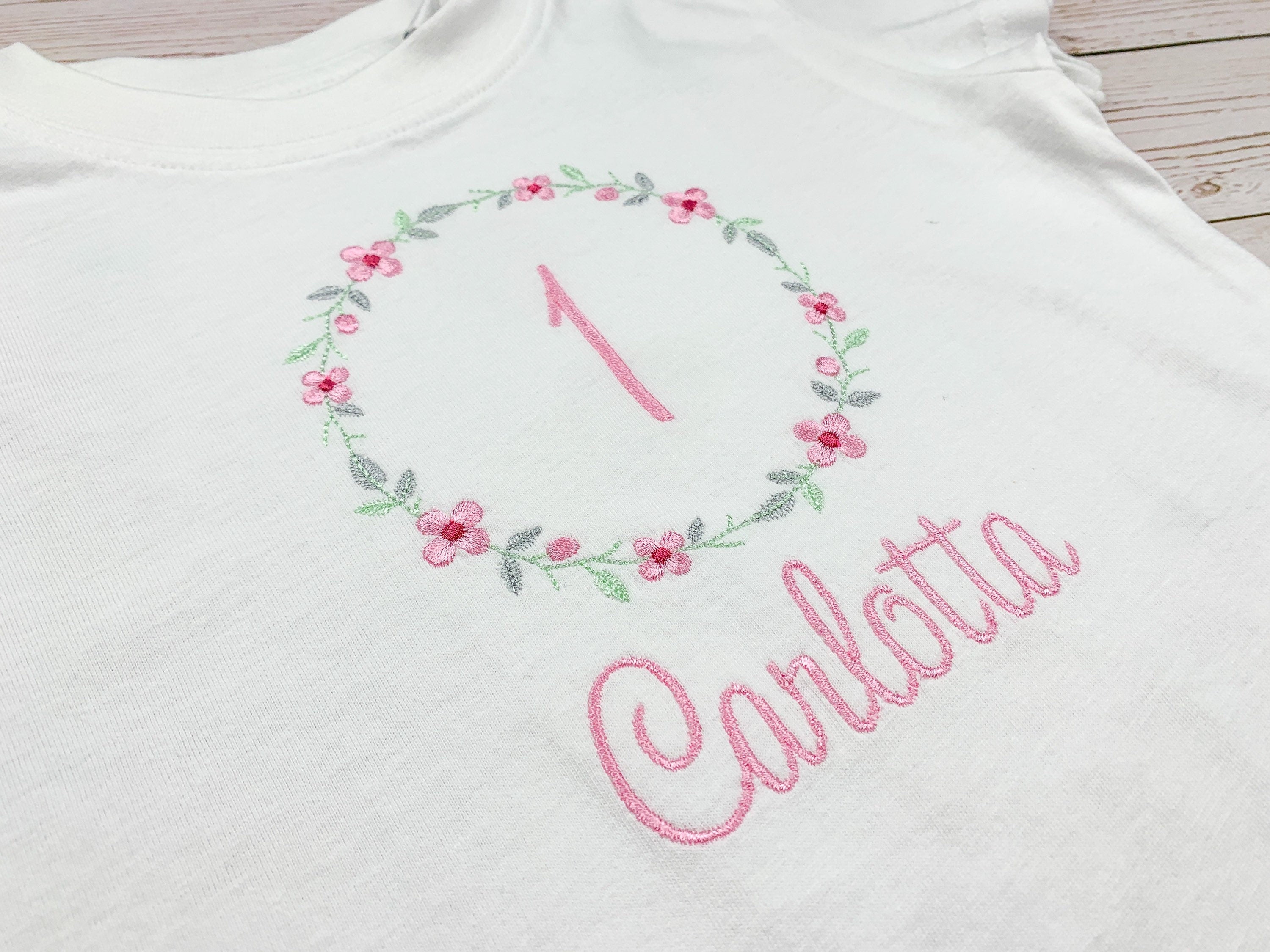 Geburtstagsshirt bestickt mit Blumenkranz und Name. Blumenkranz in den Farben mint, grau und rosa gehalten. Schrift und Zahl in rosa.