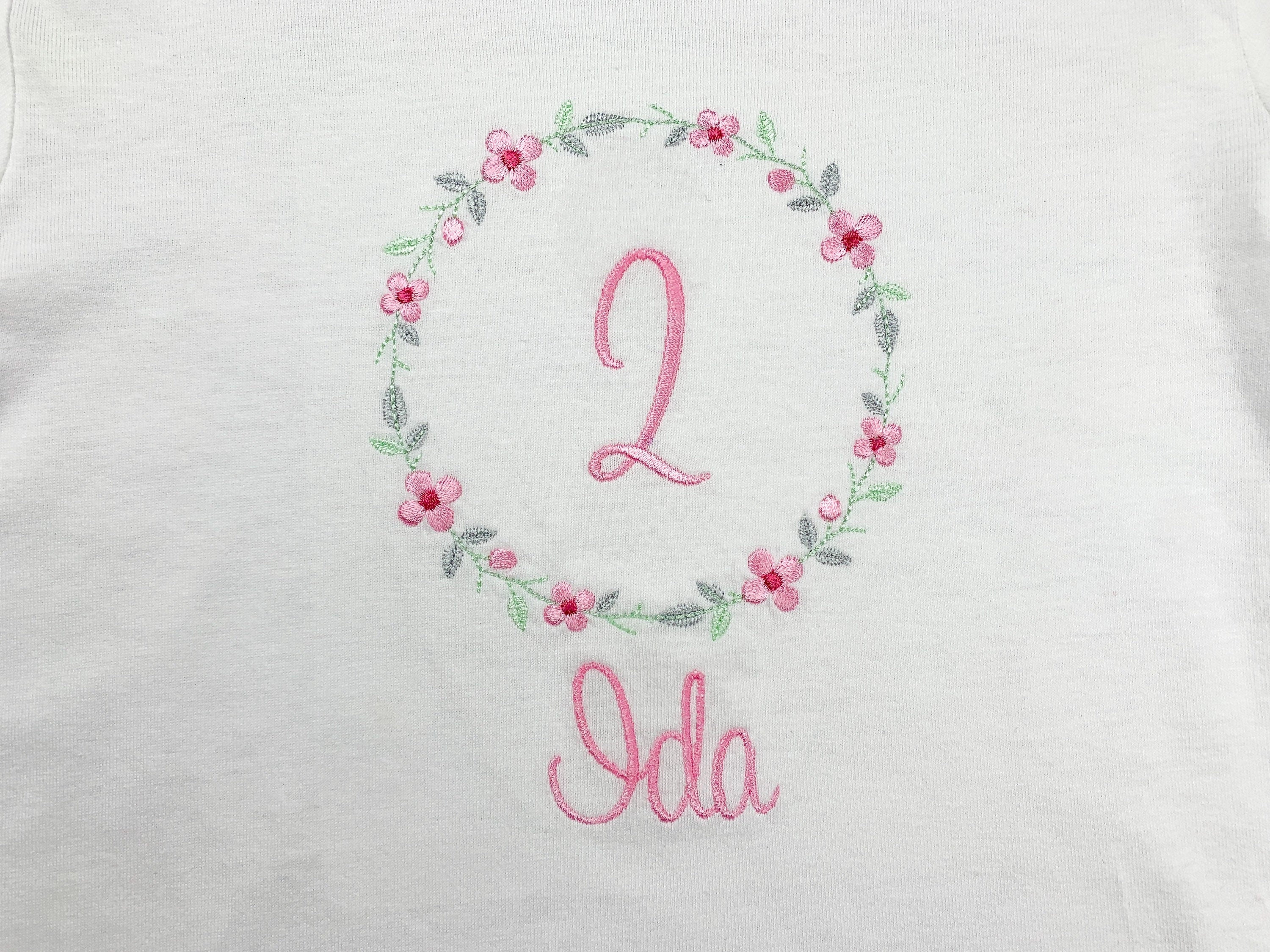 Geburtstagsshirt bestickt mit Blumenkranz und Name. Blumenkranz in den Farben mint, grau und rosa gehalten. Schrift und Zahl in rosa.