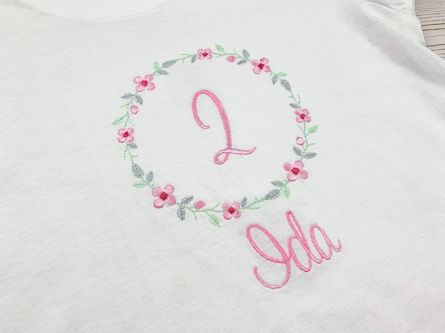 Geburtstagsshirt bestickt mit Blumenkranz und Name. Blumenkranz in den Farben mint, grau und rosa gehalten. Schrift und Zahl in rosa.