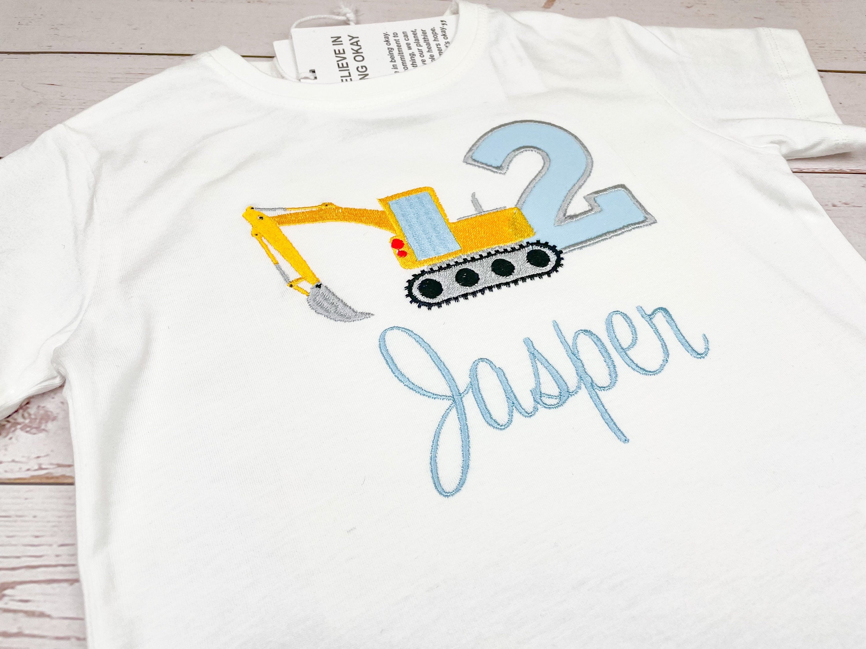 Geburtstagsshirt mit Bagger in der Farbe gelb mit großer Zahl in grau, hinterlegt mit Stoff in hellblau. Schrift Name in hellblau.