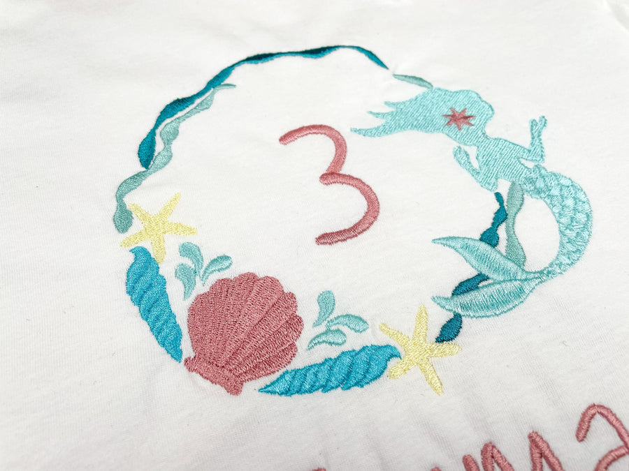  Geburtstagsshirt Meerjungfrau für Mädchen. Kreis aus Muscheln und Seegras mit Meerjungfrau in den Farben petrol, türkis und rosa.