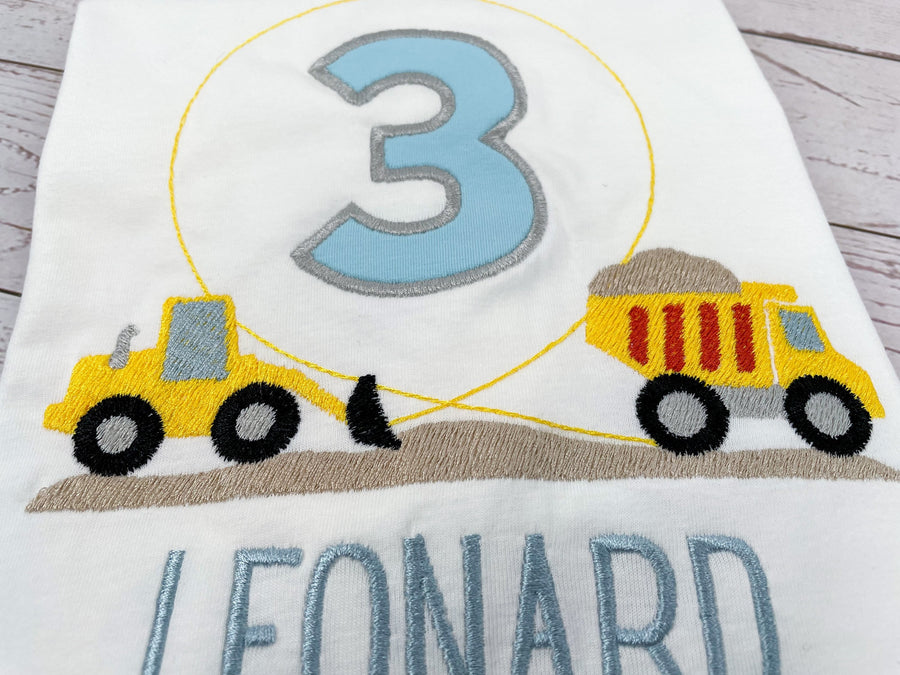 Geburtstagsshirt mit Baustellenfahrzeuen wie Lader, Kipper. Zahl in der Mitte und Schrift in hellblau.